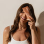 A smiling woman with long brown hair holds a tube of before Sensitive Purifying Toothpaste w/ Fluoride, Anti-Cavity in front of one eye. She wears a white tank top and stands against a plain, light-colored background.