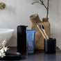A blue tube of before Whitening Purifying Toothpaste w/ Fluoride, Anti-Cavity stands on a black countertop near black containers, two bamboo toothbrushes, and a beige textured vase with a thin branch by a white sink.