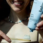 A smiling person squeezes clear Whitening Toothpaste Fluoride-Free by before from a plant-based tube onto a bamboo toothbrush. Only their mouth and hands are visible, with a brown top and beaded necklace in view.