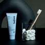 A blue tube of before Whitening Toothpaste Fluoride-Free sits next to a glass holding a bamboo toothbrush and white tablets, all arranged against a dark, minimalist background.