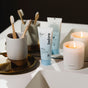 A bathroom counter features two bamboo toothbrushes in a ceramic cup, before n-HA Whitening Toothpaste Fluoride-Free, a HOMECOMING candle, a bar of soap, and another tube in the background.