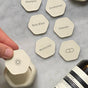 A hand holds an interchangeable Cadence magnetic label with a sun icon. Other hexagonal Cadence Labels—Set of 6—rest on marble, featuring text like shampoo and pill icons, with a bottle partially in view.