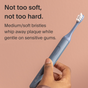 A hand holds a blue SURI electric toothbrush with 3x SURI Brush Replacement Heads against a peach background. Text reads: Not too soft, not too hard. Medium/soft bristles clean plaque gently on sensitive gums.
