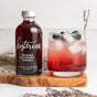 A bottle of Kosterina Crushed Blueberry Vinegar sits beside a glass of iced red drink garnished with blueberries and lavender on a wooden board, set against a blurred floral background.
