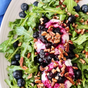 A fresh arugula salad topped with blueberries, goat cheese, Kosterina Crushed Blueberry Vinegar, and chopped pecans is served on a white plate.