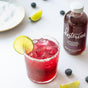 A glass of red iced drink with a salted rim and lime slice sits on a white surface next to Kosterina Crushed Blueberry Vinegar, a fruit vinegar with no added sugar, plus lime wedges, blueberries, and a plate.