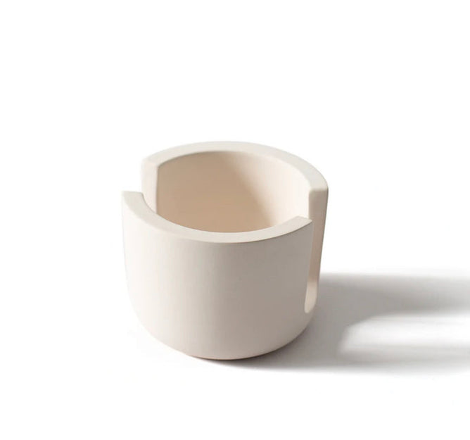 A Scrub Be Love Diatomite Sponge Holder sits next to a matte, off-white ceramic cup with two wide, symmetrical rim notches, both casting soft shadows on a white background.