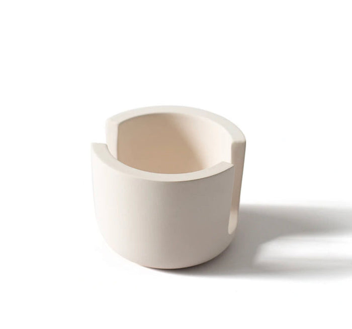 A Scrub Be Love Diatomite Sponge Holder sits next to a matte, off-white ceramic cup with two wide, symmetrical rim notches, both casting soft shadows on a white background.