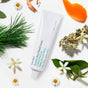 A white tube of Ursa Major Botanic Buzz Hand Cream rests on a white surface amid green leaves, orange peel, bergamot, white flowers, and pine needles—a nourishing blend designed to rehydrate and refresh your skin.