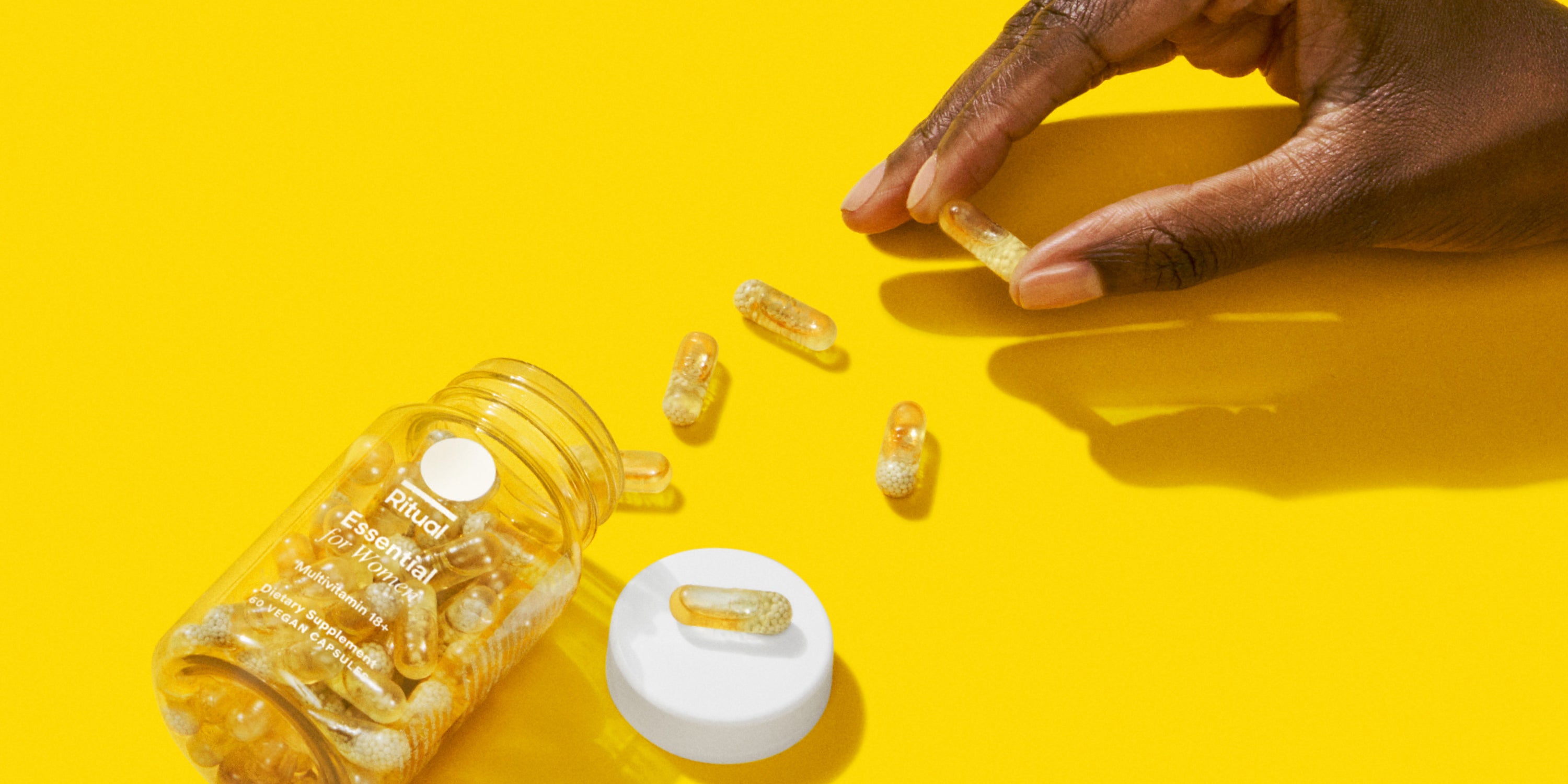 Hand holding a supplement capsule above an open bottle on a yellow background