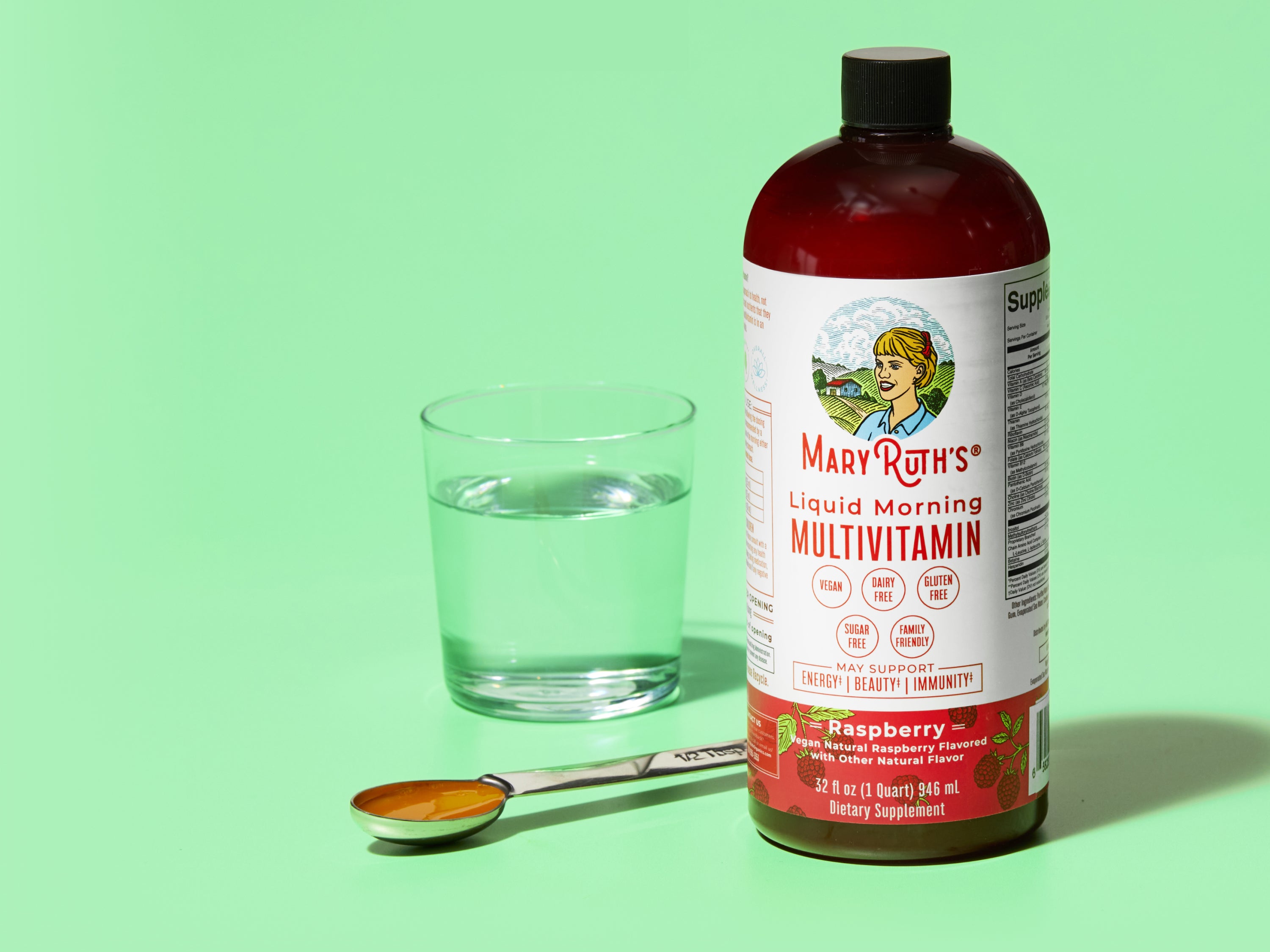 A bottle of Mary Ruths Liquid Morning Multivitamin (Raspberry flavor) stands next to a glass of water and a filled tablespoon on a green background.