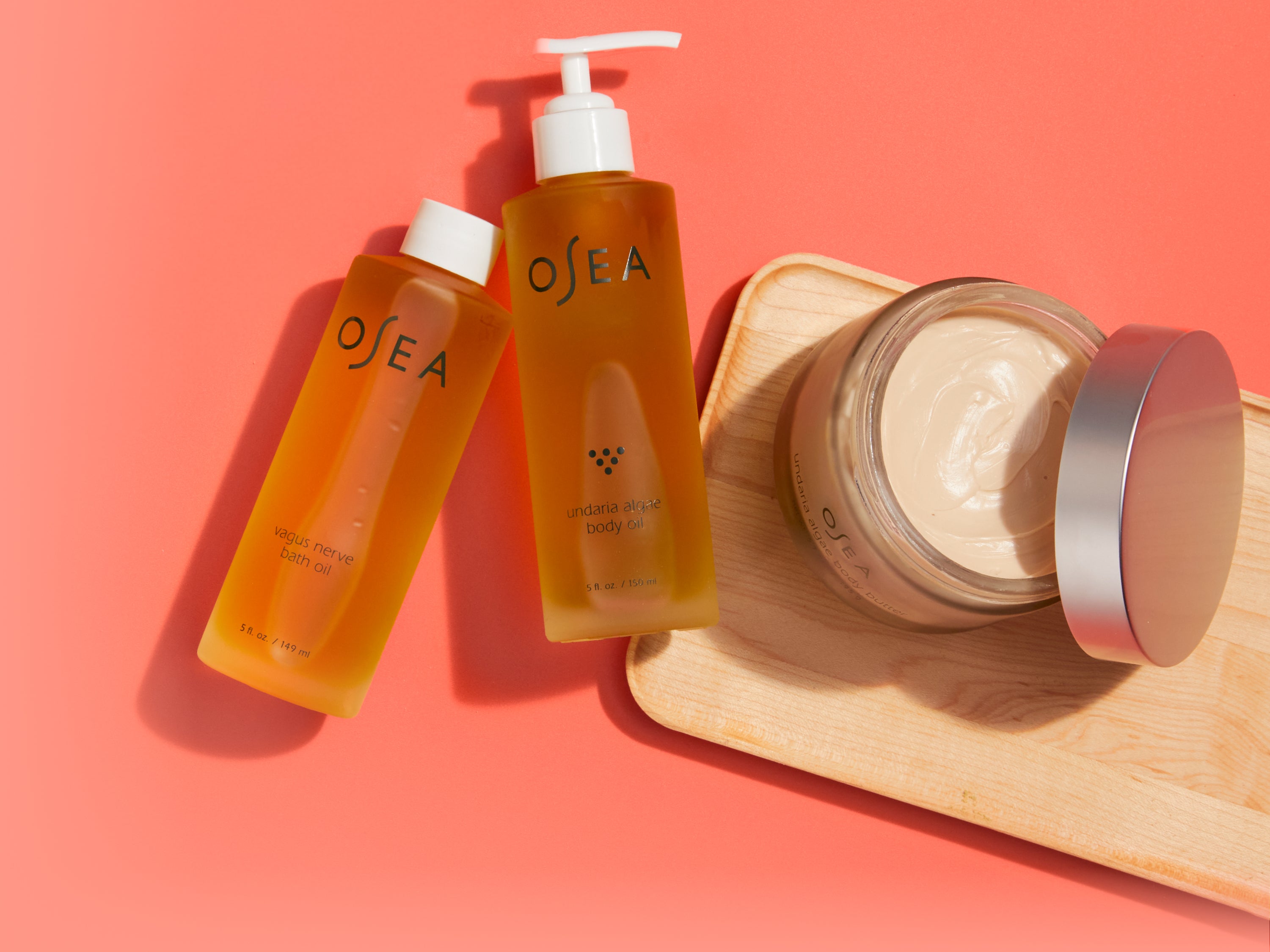 Two OSEA-branded bottles of body oil and a jar of cream sit on a wooden tray against a coral pink background. One bottle has a pump dispenser; the jar is open, showing the cream inside.