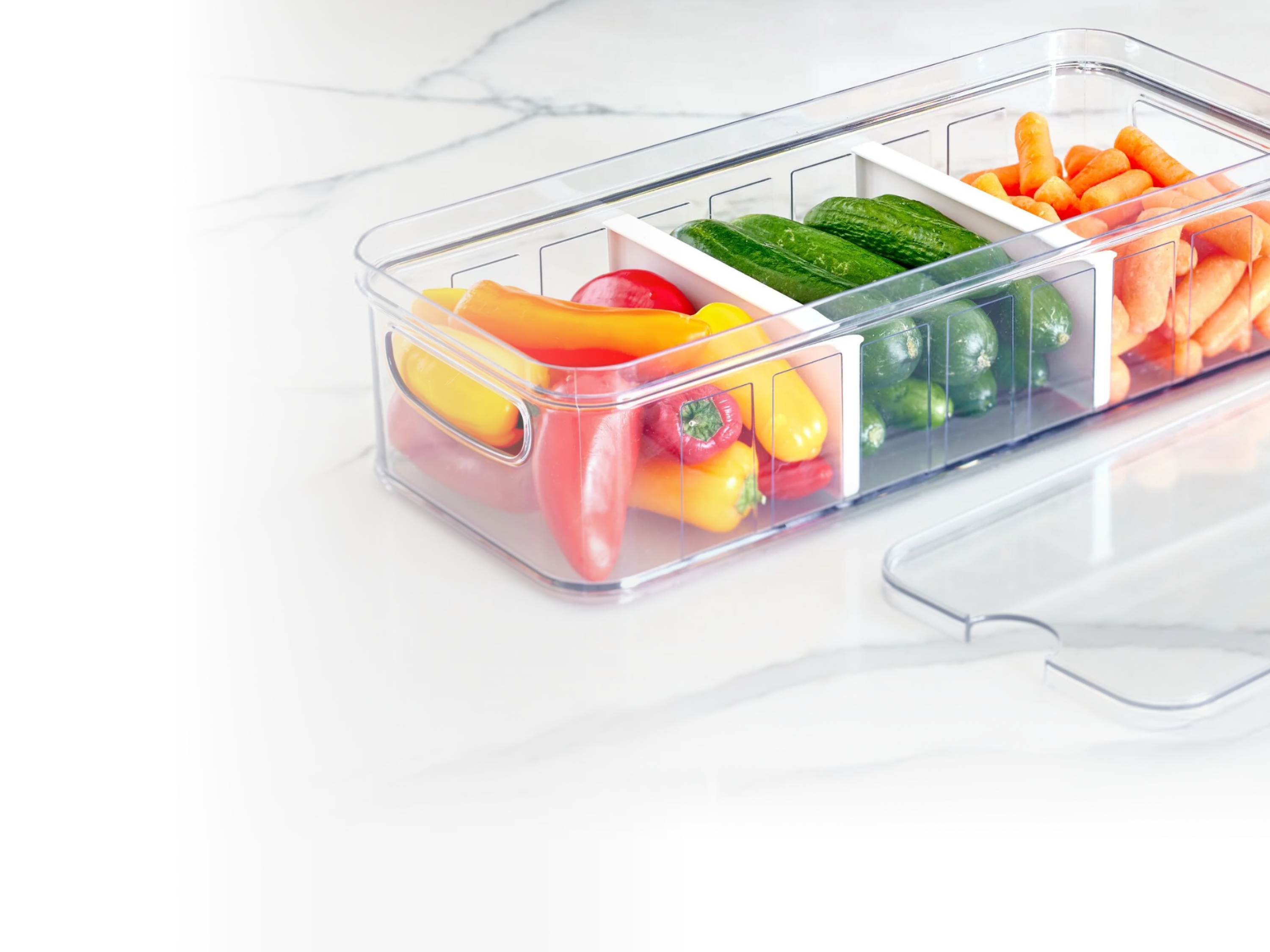 A clear plastic storage container with dividers holds mini bell peppers, cucumbers, and baby carrots. The containers lid is set beside it on a white marble surface.