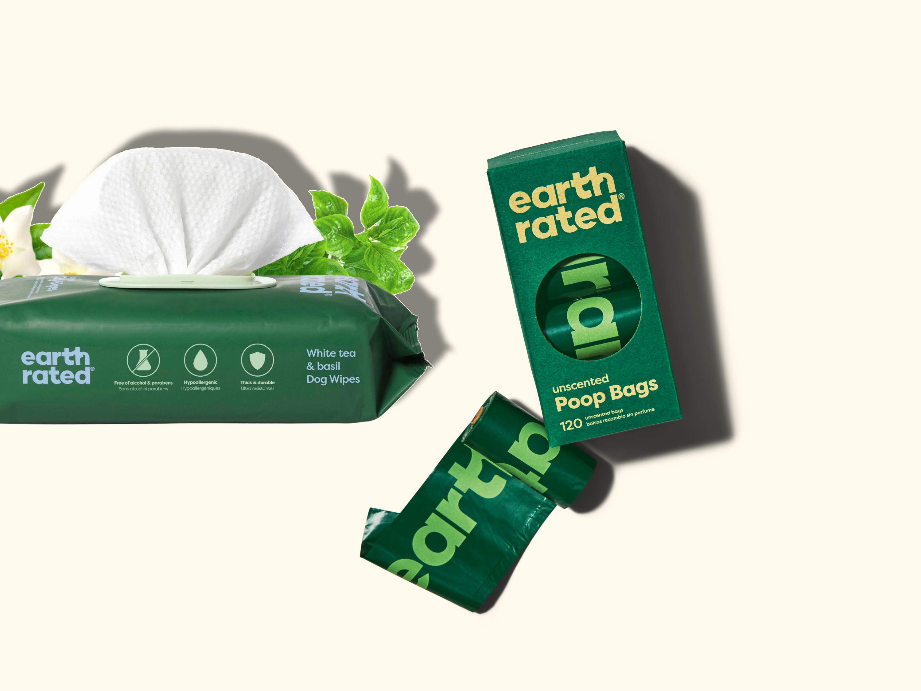 A pack of Earth Rated white tea & basil dog wipes, a box of Earth Rated unscented poop bags, and a roll of green poop bags on a beige background. Green leaves and flowers are near the wipes.