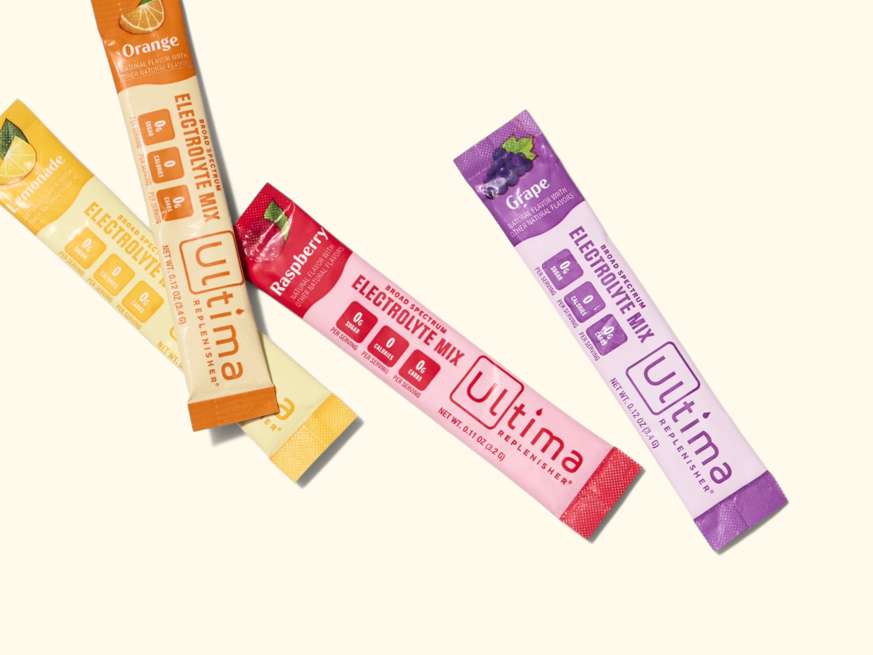 Four Ultima Replenisher electrolyte mix packets in flavors orange, lemonade, raspberry, and grape are arranged on a light background. Each stick is colorful and features icons for zero sugar, zero calories, and vegan.
