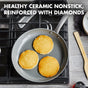 A nonstick frying pan with three golden pancakes sits on a gas stove. The background includes a wooden spatula, white plate, and dark napkin. Text reads: Healthy ceramic nonstick, reinforced with diamonds.
