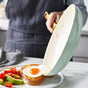 Using a light blue frying pan from the GreenPan Reserve Ceramic Nonstick 10-Piece Cookware Set, someone slides a fried egg onto toast. Nearby, a plate with avocado, cherry tomatoes, and toast rests on the marble countertop.
