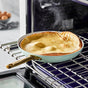 A golden-brown Dutch baby pancake bakes in a GreenPan Reserve Ceramic Nonstick skillet on an oven rack, steam rising above it. In the background, a bowl of eggs sits blurred on a light countertop.