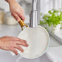 A person washes a GreenPan Reserve Ceramic Nonstick frying pan from the 10-Piece Cookware Set under a running kitchen faucet with a white sponge. Green herbs are visible by the window in the background.