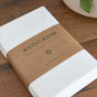 A folded Avocado Organic Cotton Crib Sheet, GOTS certified and white, is wrapped in a brown paper band and rests on a wooden surface beside a white pot with a green leaf.