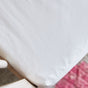 Close-up of an Avocado Organic Cotton Crib Sheet in white, neatly fitted over a crib mattress corner, with a pink and white textured rug visible on the floor nearby.