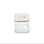 The Caraway Glass Medium Container (2.5 qt) is a clear, square, non-toxic food storage container with a beige airtight lid and central button—ideal for pantry organization. Shown empty against a white background.