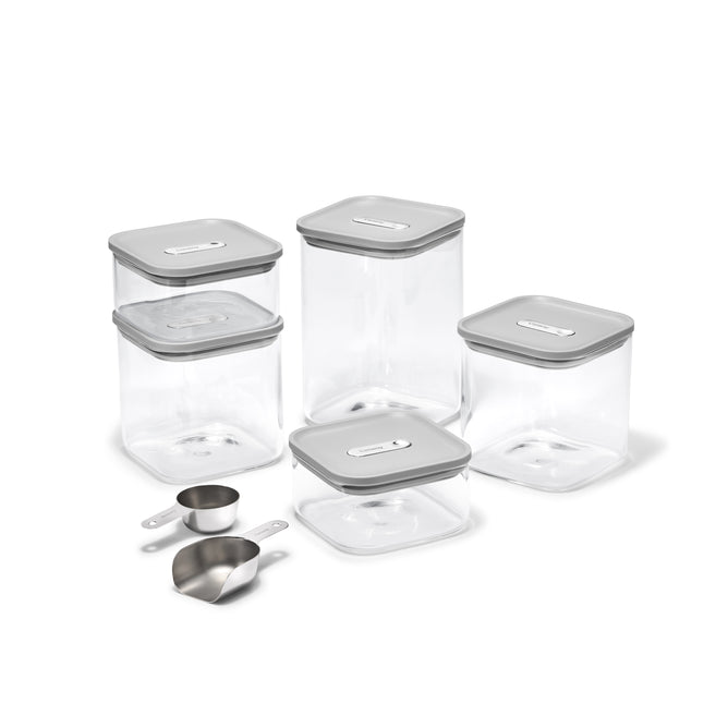 The Caraway Glass Airtight Container Set, 14-pc, features clear square containers with gray lids for pantry organization. In front are three stainless steel scoops: two measuring cups and one scoop. All containers are empty and clean.