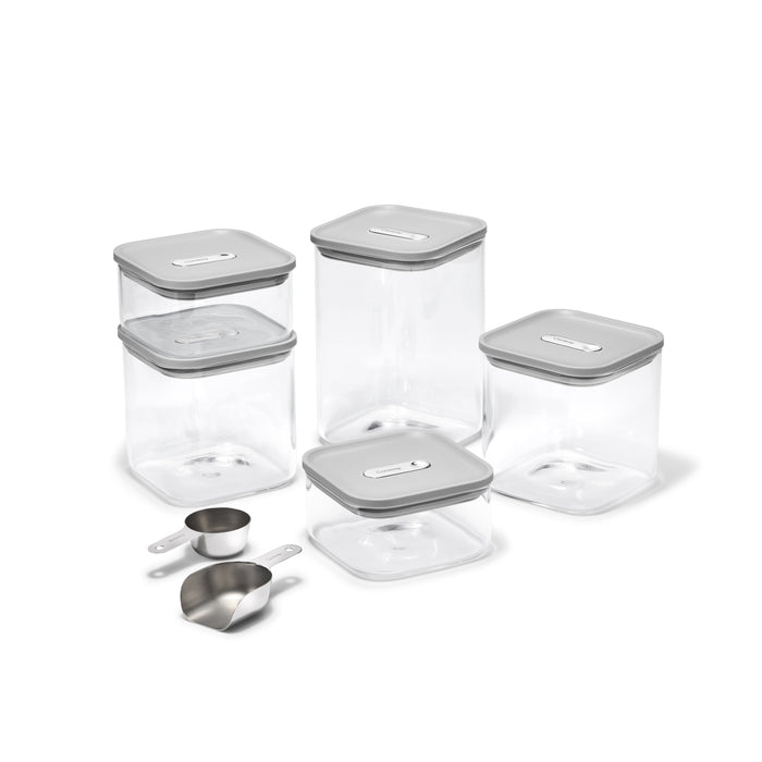 The Caraway Glass Airtight Container Set, 14-pc, features clear square containers with gray lids for pantry organization. In front are three stainless steel scoops: two measuring cups and one scoop. All containers are empty and clean.