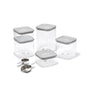 The Caraway Glass Airtight Container Set, 14-pc, features clear square containers with gray lids for pantry organization. In front are three stainless steel scoops: two measuring cups and one scoop. All containers are empty and clean.