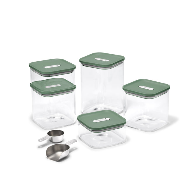 The Caraway Glass Airtight Container Set, 14-pc, features clear square containers with green lids and labels, ideal for pantry organization, displayed with two metal measuring cups and a metal scoop in front.