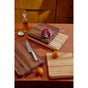 Three Caraway Edge-Grain Wood Prep & Serve Butcher Blocks (17”) rest on a red countertop—one with halved radicchio, one with a chef’s knife, and one empty. A pear, orange, oil bottle, and small bowl of salt sit nearby.