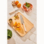 A wooden cutting board with the Caraway 5 Piece Knife Set—featuring German stainless steel chef’s knife with ergonomic handle—rests among sliced orange wedges, ginger, salad, oranges, spices, scissors, and herbs. Brand: Caraway.