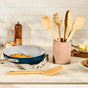 A kitchen counter displays a Caraway ceramic-coated pan with an omelet, Caraway’s 5 Piece Utensil Set in a ceramic holder, a pepper grinder, eggs in a glass bowl, butter on a dish, and bread on a cutting board against white tile.