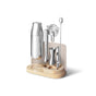 The Caraway Stainless Steel Complete Bar Set, 9-pc, includes a muddler, strainer, jigger, bar spoon, and stand—all showcased on a light wooden and white holder—perfect for at-home mixology.