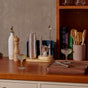 A wooden countertop featuring the Caraway Stainless Steel Complete Bar Set, 9-pc, with a pink holder, cocktail glasses with lime garnish, pepper grinder, oil dispenser, books, and wooden tray—ideal for stylish at-home mixology under warm lighting.