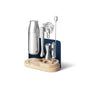 The Caraway Stainless Steel Complete Bar Set, 9-pc, includes a shaker, muddler, strainer, jigger, bar spoon, opener, and more—all elegantly displayed upright on a light wooden stand with a blue back panel. Ideal for stylish at-home mixology.