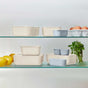 The Caraway 13 Piece Glass Food Storage Set in assorted beige and blue is neatly arranged on glass shelves, displayed with lemons, a carton of eggs, and fresh parsley.