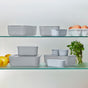 The Caraway 13 Piece Glass Food Storage Set is neatly displayed on two glass shelves, with fresh lemons, parsley, and a white bowl of eggs beside the non-toxic containers and their matching lids.