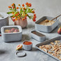 A kitchen scene features the Caraway 13 Piece Glass Food Storage Set in grey, holding strawberries, chia seeds, and oatmeal. The marble countertop also displays toasted coconut, jam, figs, and a vase with orange flowers.