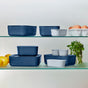 The Caraway 13 Piece Glass Food Storage Set, featuring blue and white ceramic-coated glass containers, is safely arranged on glass shelves with lemons, parsley, and eggs nearby for non-toxic food storage on a light background.