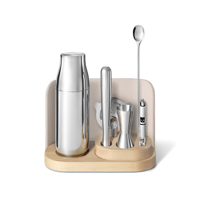 Elevate your at-home mixology with the Caraway Stainless Steel Complete Bar Set, 9-pc, featuring a shaker, muddler, strainer, jigger, spoon, and corkscrew organized on a modern light wooden and cream-colored stand.