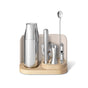 Elevate your at-home mixology with the Caraway Stainless Steel Complete Bar Set, 9-pc, featuring a shaker, muddler, strainer, jigger, spoon, and corkscrew organized on a modern light wooden and cream-colored stand.