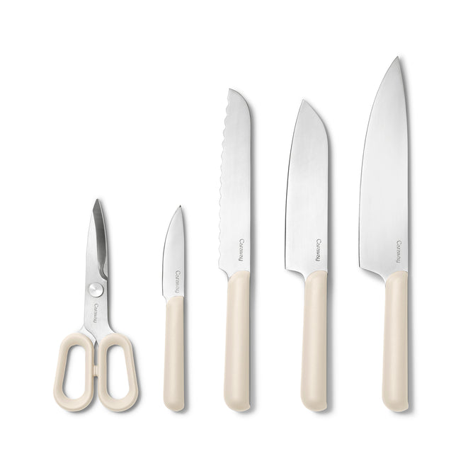 The Caraway 5 Piece Knife Set features German stainless steel kitchen scissors, a paring knife, serrated bread knife, santoku, and chef’s knife with cream ergonomic handles—arranged upright against a white background.