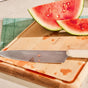 A Caraway 5 Piece Knife Set chef’s knife with an ergonomic white handle rests on a wooden cutting board with watermelon juice and slices, while a green mat appears partially in the background.