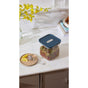 The Caraway Glass Medium Container, 2.5 qt, with its airtight blue lid, stores loose tea leaves on a marble countertop. Paired with a tea infuser and lemon slices on a beige tray, it offers non-toxic storage and stylish pantry organization.