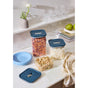 Caraway’s 14-pc Glass Airtight Container Set with blue lids holds cereal, coconut flakes, and pistachios on a white countertop, surrounded by blue plates, green apples, glassware, and a vase—stylish pantry organization.