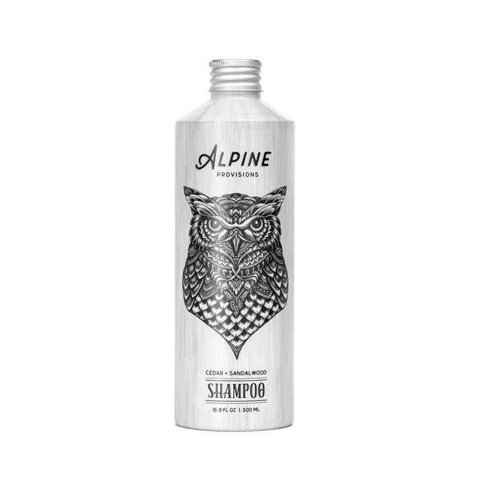 A silver Alpine Provisions Shampoo bottle, featuring a detailed black owl illustration and labeled “Cedar + Sandalwood,” 16.9 fl oz (500 mL), highlights natural hair care with essential oil aromatherapy on a white background.