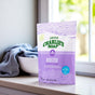 A bag of Charlie's Soap Laundry Booster & Hard Water Treatment Powder sits on a windowsill beside a white bowl filled with light blue laundry, as sunlight brightens the clean, minimal setting.