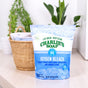A bag of Charlie’s Soap Oxygen Bleach Powder, a chlorine-free laundry booster and stain remover, sits on a light wooden surface with a wicker basket of folded towels and green potted plants in the background.