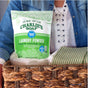 A person in a denim shirt holds a woven basket with Charlie's Soap Powder Laundry Detergent - Ultra Concentrated, a white towel, and a green-striped cloth.